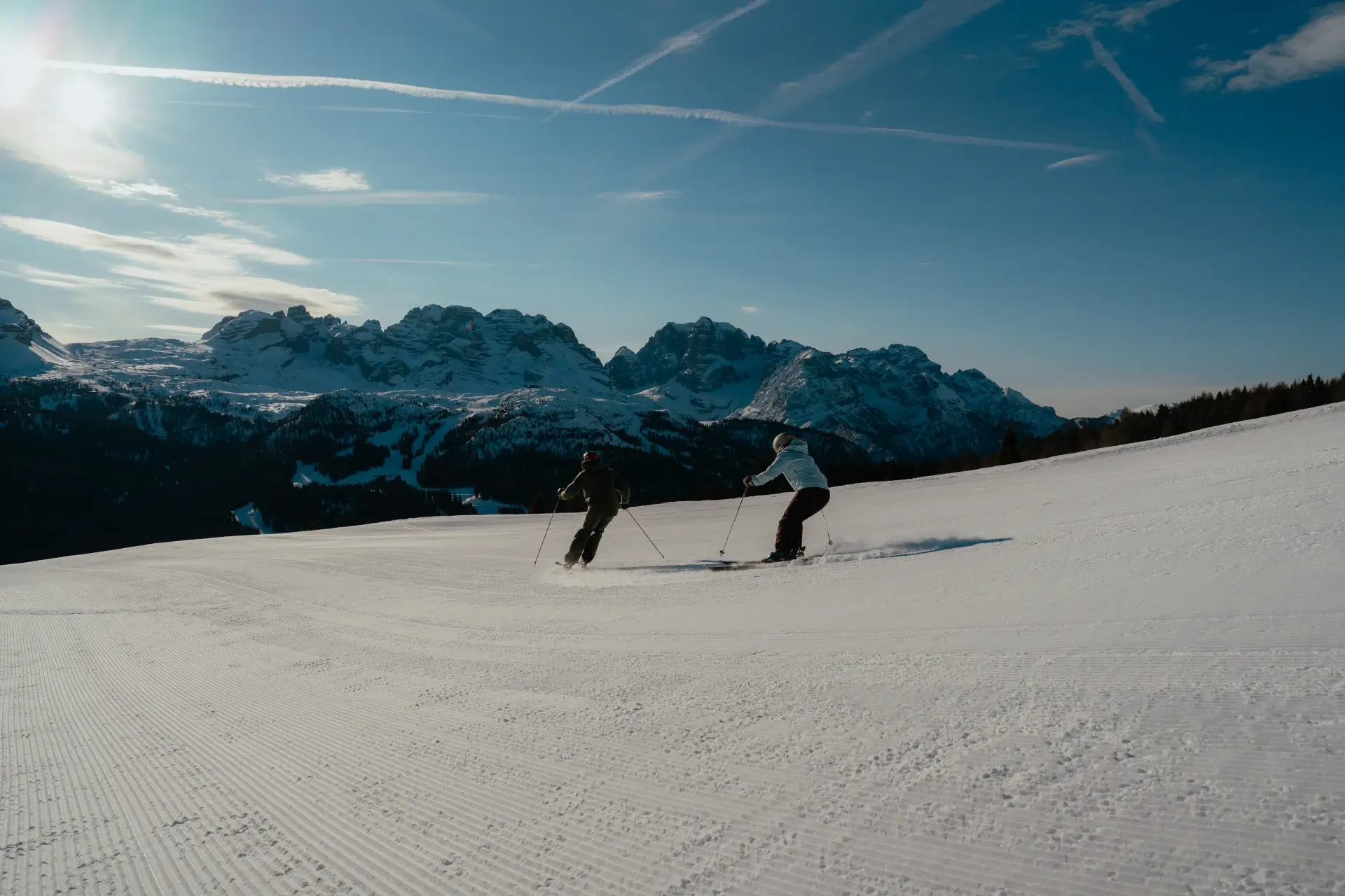 Due persone sciano su una pista innevata con montagne sullo sfondo.